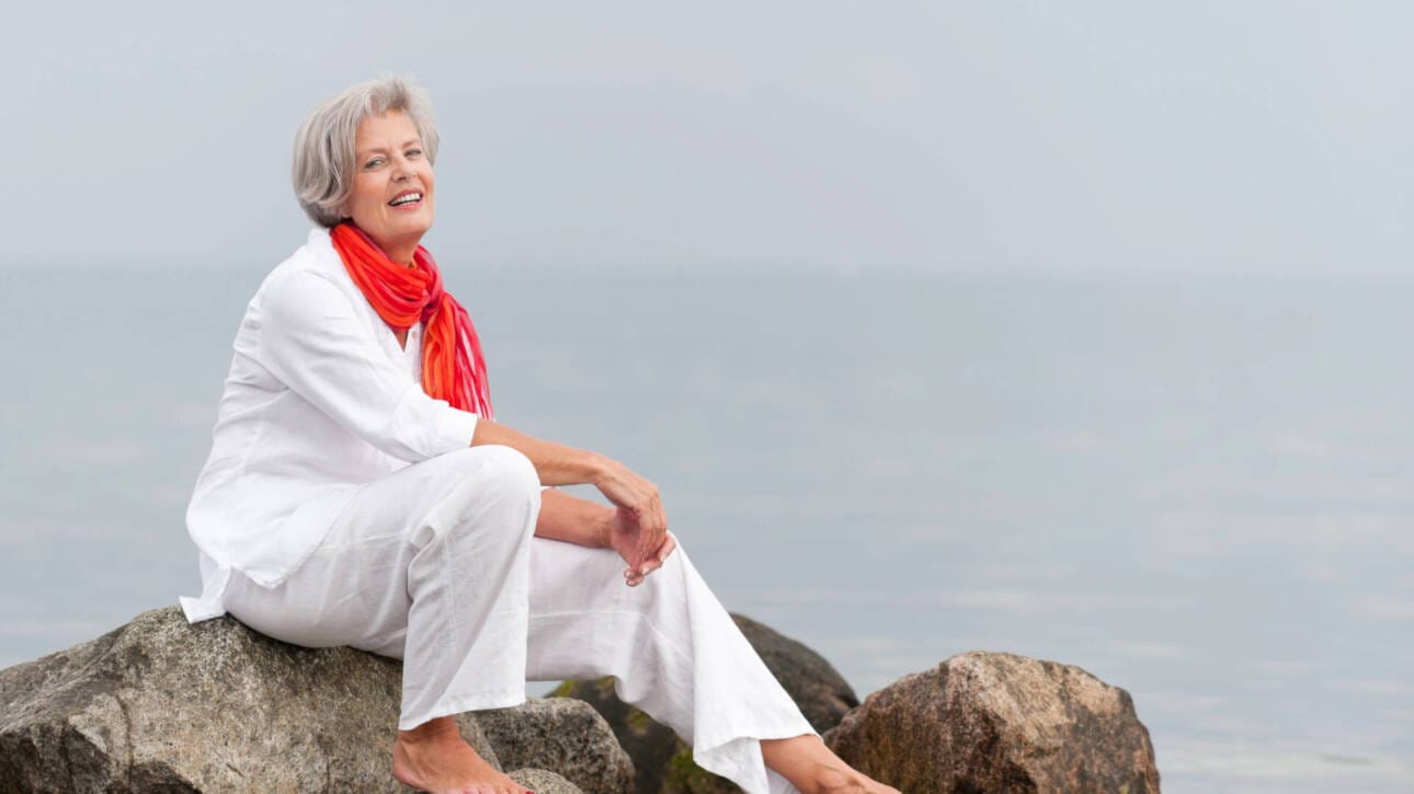 Symbolbild Frau am Strand sitzend auf Felsen. Monatsthema „Gesund alt werden: einfachSchritte“ für ViewMed|Depositphotos @PicterArt|Lizenznehmer: Pressebüro LAAKS