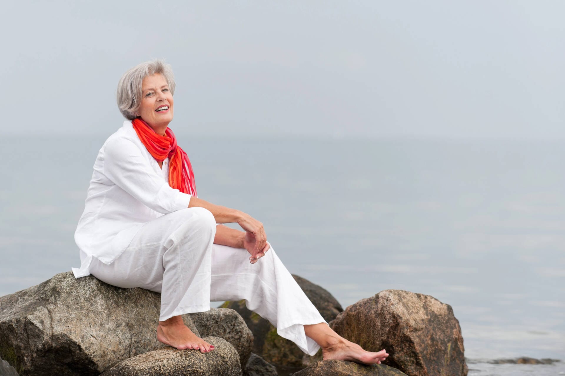 Symbolbild Frau am Strand sitzend auf Felsen. Monatsthema „Gesund alt werden: einfachSchritte“ für ViewMed|Depositphotos @PicterArt|Lizenznehmer: Pressebüro LAAKS